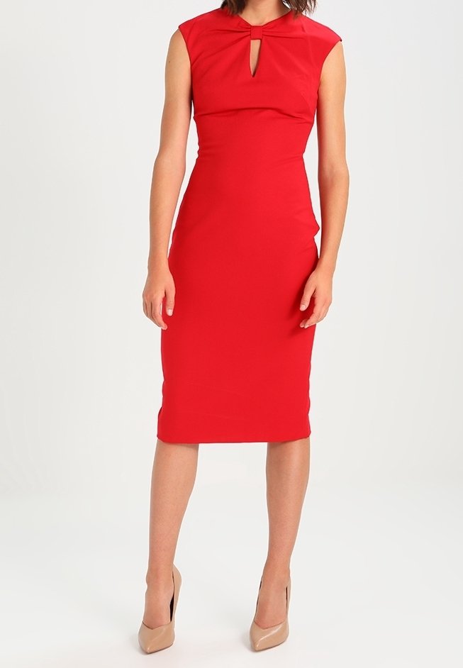 Femme portant une robe rouge moulante sans manches avec un décolleté en forme de goutte et des talons hauts beige, debout devant un fond blanc uni.