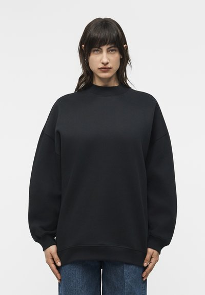 Femme aux cheveux raides et foncés portant un sweat-shirt noir oversize et un jean bleu, debout devant un fond clair uni.