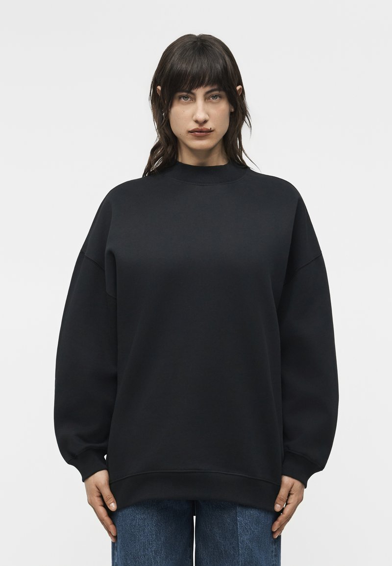 Γυναίκα με σκούρα ίσια μαλλιά, φοράει υπερμεγέθη μαύρο φούτερ και μπλε τζιν, στέκεται μπροστά σε απλό φωτεινό φόντο.