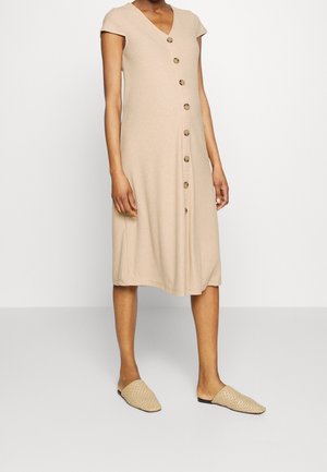 Frau trägt ein beigefarbenes, geripptes Midi-Kleid mit kurzen Ärmeln und vorderen Knöpfen, kombiniert mit gewebten beigen Slip-on-Schuhen.