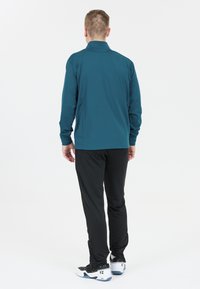 Man med ryggen mot kameran, iklädd en teal långärmad tröja, svarta byxor och vit-svarta sneakers mot en enfärgad vit bakgrund.