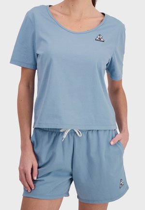 Kvinde iført lyseblå top med korte ærmer og matchende shorts med snøre og logopatche på brystet og venstre ben.