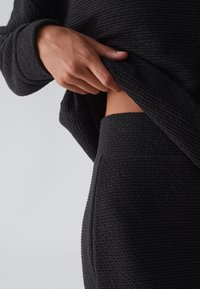 Zwart textiel met een losse pullover en goed passende broek. De hand tilt de pullover op om de tailleband van de broek te onthullen.