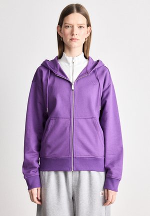 Fialová zipsová mikina vyrobená z jemnej tkaniny s prednými vreckami a kapucňou na šnúrku. V páre so svetlosivými teplákmi. Minimalistické logo na hrudi.