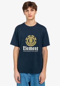 Camiseta de algodón azul marino con un logotipo circular dorado y texto blanco que dice "Elemento viento:agua:fuego:tierra". Manga corta y corte relajado.