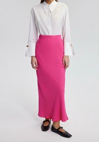 Weißes Hemd mit Kragen und langen Ärmeln, verziert mit goldenen Knöpfen, kombiniert mit einem pinkfarbenen, strukturierten Rock bis zum Knöchel. Schwarze flache Schuhe runden das Outfit ab.