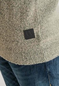 Strickpullover in Hellgrün mit gesprenkelter Textur. Mit einem schwarzen Etikett am Saum. Kombiniert mit blauen Jeans, die die Nähte der Gesäßtaschen zeigen.