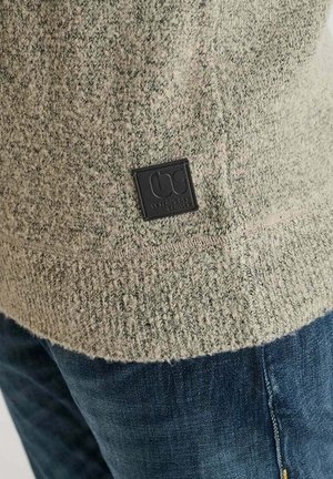 Brede gebreide trui in lichtgroen met een gespikkelde textuur. Heeft een zwart label aan de zoom. Gecombineerd met blauwe jeans met zichtbaar stiksel op de achterzak.