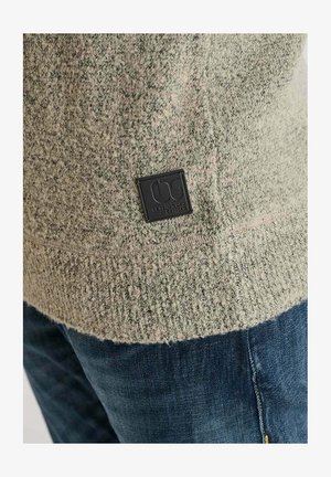 Brede gebreide trui in lichtgroen met een gespikkelde textuur. Heeft een zwart label aan de zoom. Gecombineerd met blauwe jeans met zichtbaar stiksel op de achterzak.