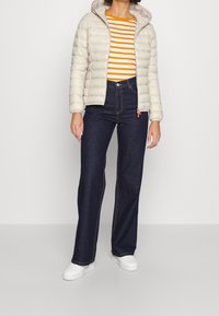 Beige gepolsterte Jacke mit Kapuze, orange-weißer gestreifter Shirt, hochgeschnittene dunkelblaue Jeans, weiße Sneakers und auffällige Nähte.