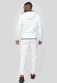 Survêtement de sport blanc avec capuche, présentant des accents et des zips noirs, au design ajusté et en tissu texturé, porté avec des chaussures noires et orange.