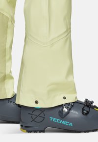 Ljusgula skidbyxor som täcker mörkgrå Tecnica skidstövlar med turkosa logotyper och spännen på en vit bakgrund.