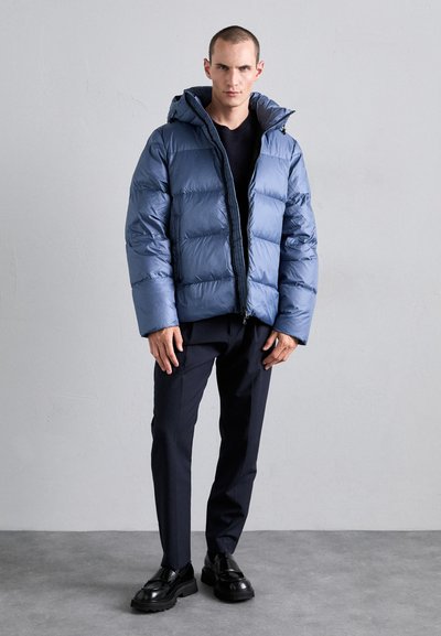 Mattblaues Pufferjackett aus leichtem Stoff mit Kapuze und gestepptem Design. Kombiniert mit dunklen Hosen und schwarzen Schuhen mit strukturierten Sohlen.