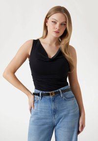 Top nero senza maniche con scollatura a cowl, abbinato a jeans a vita alta di un blu chiaro e una cintura nera con fibbia dorata a forma di lettera.