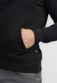 Sudadera negra de mezcla de algodón, con bolsillo canguro, puños y dobladillo acanalados. Pequeña etiqueta "Petrol" cerca del bolsillo.