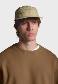 Beige Kappe mit gesticktem Elefantenlogo, verfügt über eine weiche Textur und ein strukturiertes Design. Darunter ein brauner Pullover mit besticktem Elefantendetail.