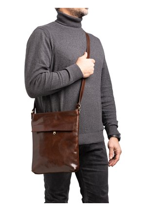 Homme portant un pull à col roulé gris et un pantalon noir, portant un sac bandoulière en cuir marron avec un rabat et une fermeture.