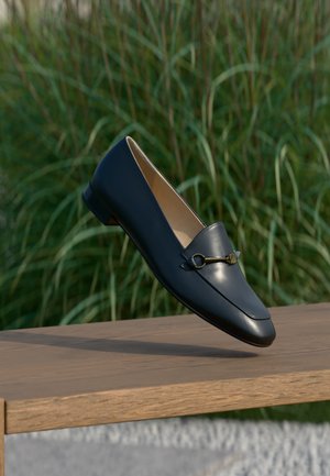 Schwarzer Lederloafer mit goldenem Horsebit-Detail schwebt über einer hölzernen Bank, grüner Gras-Hintergrund.