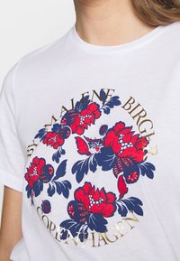 Camiseta de algodón blanca con un diseño floral circular en rojo y azul, con texto dorado que dice "By Malene Birger" y "Copenhague."
