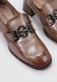 Chaussures en cuir marron et brillant avec un bout carré, présentant un accent de chaîne noire sur le dessus et un talon carré. Fini texturé visible.