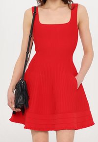 Femme portant une robe rouge à manches courtes avec un décolleté carré, portant un sac bandoulière à franges noir, la main dans la poche de la robe.