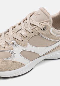 Beige og hvite sneakers med semsket skinn og mesh-paneler, dekorative sølvfarget aksenter og en teksturert såle med snøring.