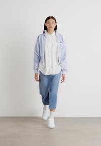 Šviesiai mėlynas bomber stiliaus švarkas su elastingomis rankovėmis, po juo balta marškinėlių chemise, bei šviesiai mėlyni laisvi džinsai. Balti sportbačiai ir vientisas fonas.