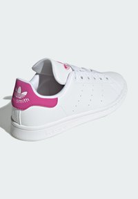White leather sneaker with a smooth texture, pink accent on the heel, perforated detailing on the sides, and flat rubber sole.