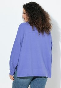 Mujer con cabello rizado, vistiendo una blusa morada de manga larga y suelta, y jeans azules, de perfil contra un fondo blanco liso.