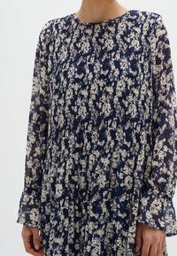 Robe florale bleu marine avec des manches longues plissées ; présente un col rond et des motifs floraux de couleur claire dispersés sur l'ensemble.