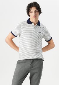 White and navy striped polo shirt with a collared neckline, short sleeves, and embroidered logo. Worn with gray trousers.