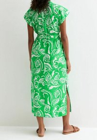 Vihreä kukkamekko, jossa on valkoisia lehtikuvioita, lyhyet hihat ja sivuhalkio. Valmistettu kevyestä kankaasta, joka näyttää rentoa istuvuutta.