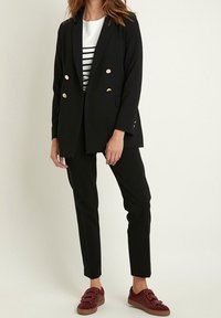 Černý přizpůsobený sako se zlatými knoflíky, doplněné o černé slim-fit kalhoty a burgundské tenisky. Pod tím je vidět pruhované bílé tričko.