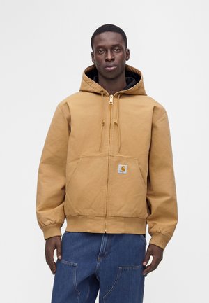 Mand iført en beige lynlås hættetrøje og blå jeans, der står mod en ensfarvet lys baggrund.