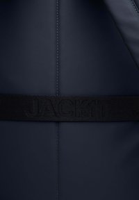 Giacca imbottita blu navy con una texture liscia, dotata di una cintura in tessuto nera etichettata "JACKIT" e un dettaglio con cucitura centrale.