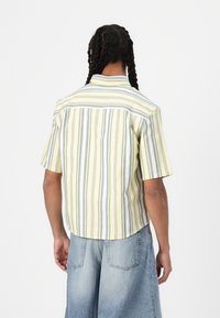 Camicia a maniche corte gialla con righe grigie, realizzata in tessuto leggero. Presenta un orlo curvo e un colletto standard.