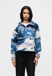 Γυναίκα που φοράει μπλε και λευκή φλις μπλούζα με μοτίβο και ψηλό γιακά, καθώς και μαύρο παντελόνι, στέκεται μπροστά σε απλό φόντο.