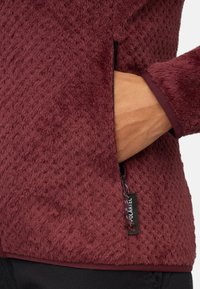 Giacca in pile bordeaux con superficie testurizzata, tasca laterale con cerniera e etichetta con marchio. I polsini sono lisci e aderenti.