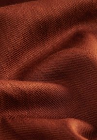 Tissu tricoté doux de couleur rouille avec une texture de tissage visible et des plis naturels créant des variations de lumière et d’ombre.