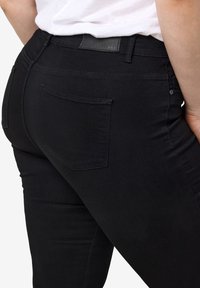 Gros plan d'une personne portant un jean noir ajusté avec des poches arrière et une chemise blanche rentrée.