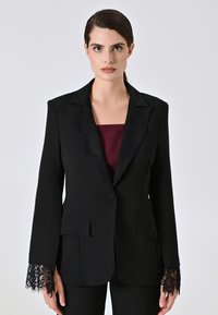 Blazer nero con dettagli in pizzo sulle maniche e sul rever, dotato di chiusura a un bottone e due tasche frontali, indossato su un top bordeaux.