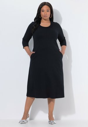 Svart klänning med långa ärmar, rundad halsringning och sidofickor, med rak skärning och slät textur, bärs med metalliska silverfärgade skor.