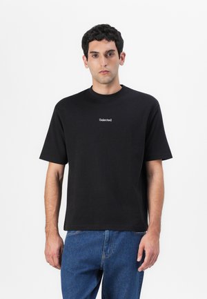 Black cotton t-shirt with a round neck and short sleeves, featuring a white "Selected" logo in the center. Worn with blue jeans.
