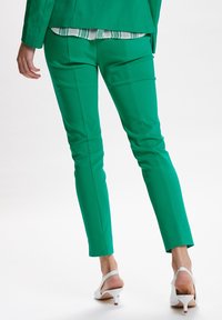 Zielone dopasowane spodnie, slim fit, gładka tekstura, z fałdami z przodu. Noszone z białymi sandałami na obcasie, eksponując warstwowy zielono-biały top.