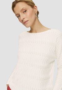 Pull en tricot côtelé blanc avec une texture ondulée, col rond et manches longues. Le tissu présente des détails de ventilation subtils.