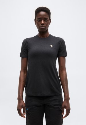 Kvinde med kort hår iført en tætsiddende mørkegrå kortærmet skjorte og sorte bukser, stående mod en ensfarvet lys baggrund.