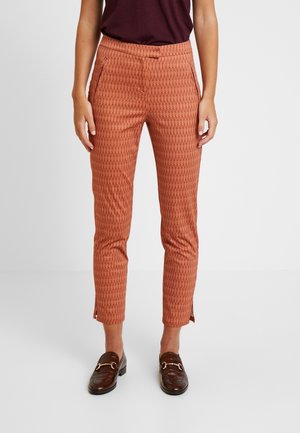 Mujer con pantalones pitillo hasta el tobillo de color óxido con estampado y aberturas laterales, combinados con mocasines de cuero marrón y una blusa oscura.