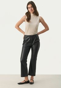 Beige ärmelloses Oberteil, kombiniert mit hochgeschnittenen schwarzen Hose aus Kunstleder, die eine glatte Textur und einen subtilen Glanz aufweist. Schwarze flache Schuhe runden den Look ab.