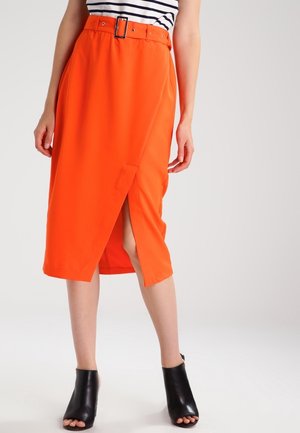 Gonna midi arancione con vita regolabile, spacco laterale e texture liscia. Indossata con scarpe nere aperte.