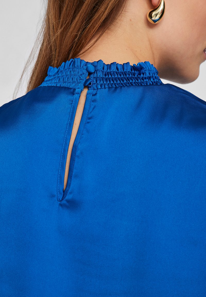 Blauwe satijnen blouse met een smockhalslijn en een knoopsluiting aan de achterkant. De stof heeft een soepele textuur met een lichte glans.